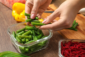 Woman putting green beans into glass container at wooden table, closeup. Food storage