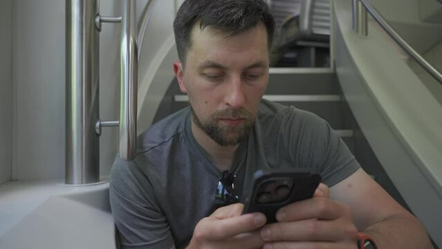 Young man travels on double decker train and sits on flyer using his smartphone. No available seating during railroad trip. Male commutes in dual deck train seated on flyer and surfing internet. 