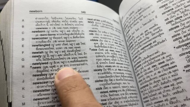 Hand open English - Thai dictionary for reading. Close-up macro shot looking through a book turning the pages. Business and education concept. Pointing at word "News"