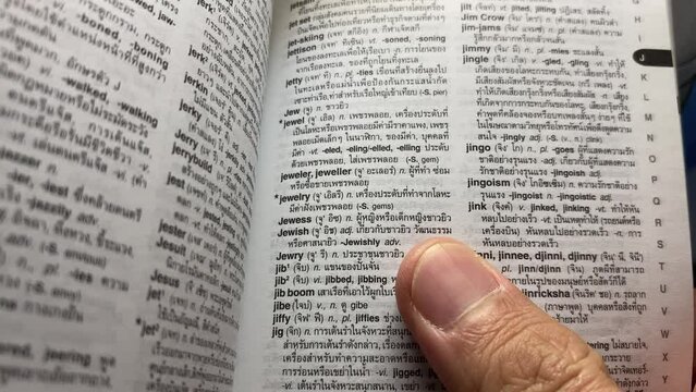 Hand open English - Thai dictionary for reading. Close-up macro shot looking through a book turning the pages. Business and education concept. Pointing at word "Jewish"