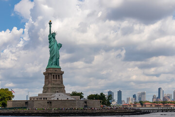 Fototapeta premium Iconic representation of freedom and independence, the Statue of Liberty with flaming torch on Liberty Island. The Lady on a Pedestal is surrounded by clouds. New York City visible in the back.