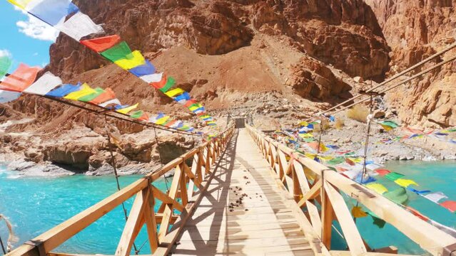 4K shot of wooden suspension bridge over a clear blue Tsarap river surrounded by mountains in Zanskar, Ladakh, India. Buddhist prayer flags on wooden bridge near Phugtal monastery. 