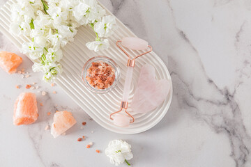 A rose quartz cosmetic roller massager, a gua sha scraper and flowers lie on a white podium in the...