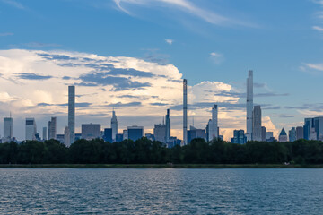Captivating New York urban skyline at dusk with striking and modern skyscrapers reflecting on water seen from Jacqueline Kennedy Oasis Reservoir. The buildings are taller than surrounding clouds.