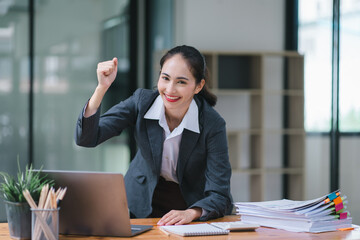 Portrait of a happy young businesswoman celebrating success with arms raised in front of a laptop,...