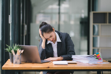 The stressed and exhausted millennial Asian businesswoman is seen sitting at her office desk with her hand on her head, indicating a hard working day where she is overloaded with work.