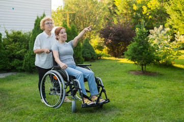 An elderly woman is carrying an adult daughter sitting in a wheelchair. Caucasian woman pointing her finger admiringly while walking outdoors. 