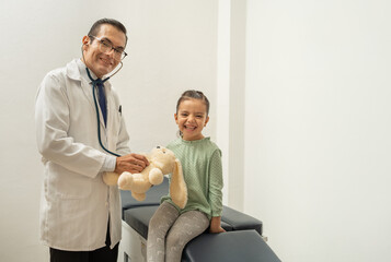 doctor using stethoscope listening teddy bear heartbeat. pediatrician building confidence on child. kid smiling at doctor's office. Doctor and girl smiling looking at camera