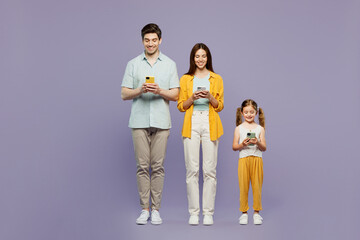 Full body young fun parents mom dad with child kid daughter girl 6 years old wear blue yellow casual clothes hold in hand use mobile cell phone isolated on plain purple background. Family day concept.