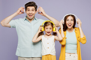 Young fun surprised parents mom dad with child kid daughter girl 6 years old wear blue yellow casual clothes listen to music in headphones dance isolated on plain purple background Family day concept