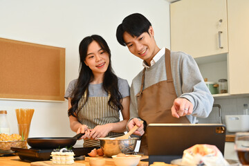 Young happy couple preparing breakfast in the kitchen at home and reading recipes on digital tablet
