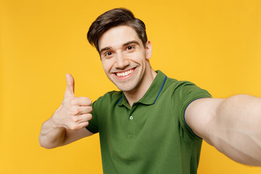 Close up young smiling happy fun man wears green t-shirt casual clothes doing selfie shot pov on mobile cell phone show thumb up isolated on plain yellow background studio portrait. Lifestyle concept.