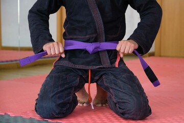 Cropped shot of an unrecognizable man tying a purple Brazilian Jiu-Jitsu belt. BJJ training.