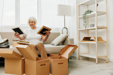 elderly woman sits on a sofa at home with boxes. collecting things with memories albums with photos and photo frames moving to a new place cleaning things and a happy smile. Lifestyle retirement.