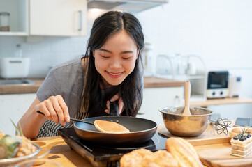 A happy Asian woman flipping a pancake in a pan with a spatula, enjoys cooking in the kitchen - obrazy, fototapety, plakaty