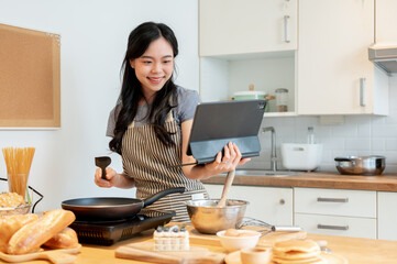 A happy Asian woman is reading an online recipe on her tablet and enjoying making pancakes