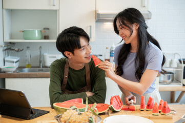 Obraz na płótnie Canvas A happy Asian man is being fed a slice of watermelon by his girlfriend in the kitchen