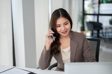 Obraz premium A beautiful Asian businesswoman is looking at her computer screen while talking on the phone