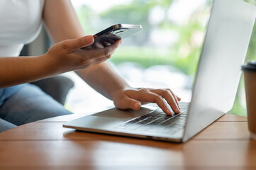 A female freelancer working remotely at a cafe, using her smartphone and laptop at a table.
