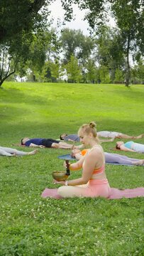 Vertical Screen: woman playing singing bowls during meditation while practitioners lie on grass in corpse pose. People resting in Shavasana, yoga class deep restoration, body and mind relaxation