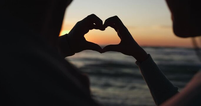 Silhouette, sunset and hands of couple with heart shape at the beach as love on holiday or tropical vacation together. Ocean, sign and people with care, support or trust symbol for hope on an island