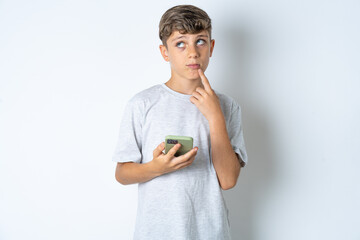 Beautiful kid boy wearing grey casual t-shirt thinks deeply about something, uses modern mobile...