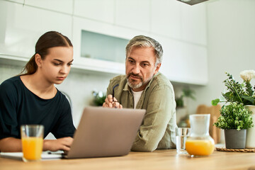 A senior man with his granddaughter using a laptop in the kitchen, both looking serious.