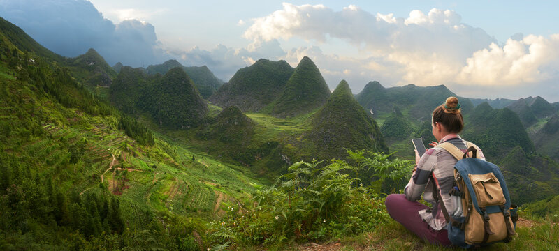 Woman Traveler with backpack resting and photographing scenic panoramic view of green mountains.
