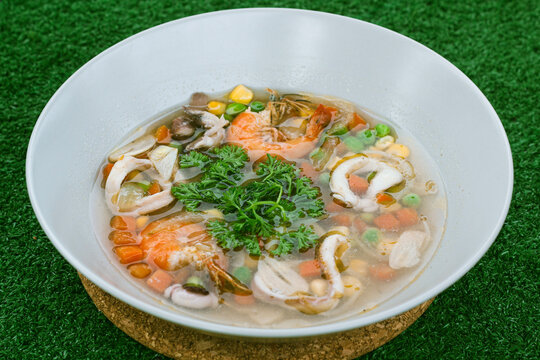 seafood soup in white bowls, soup containing pieces of ring squid, prawns, peas, carrots topped with green celery leaves and lemon slices in it, served on a gray floor.
