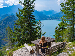 A teenage girl sits on a bench on a mountain against the backdrop of Lake Attersee, Austria and looks into the distance