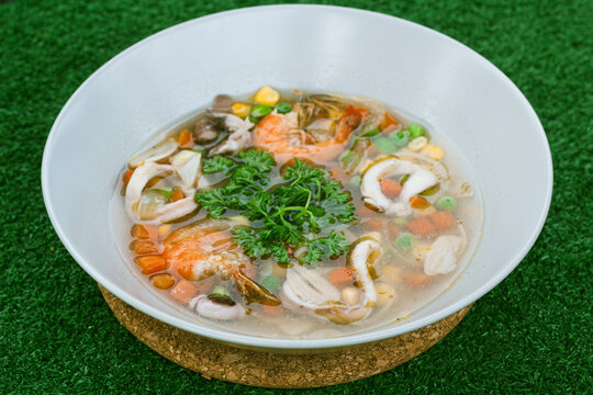 seafood soup in white bowls, soup containing pieces of ring squid, prawns, peas, carrots topped with green celery leaves and lemon slices in it, served on a green floor.