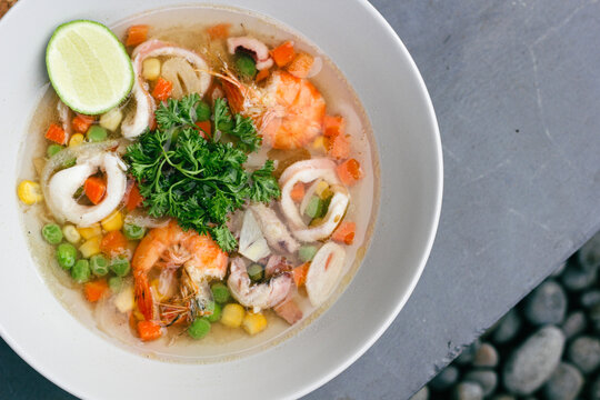seafood soup in white bowls, soup containing pieces of ring squid, prawns, peas, carrots topped with green celery leaves and lemon slices in it, served on a gray floor.
