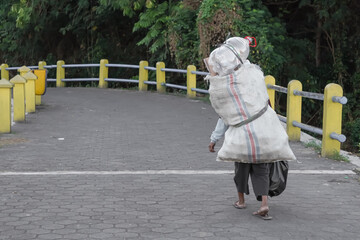 A used goods scavenger or junk collector carrying large bag on the back crosses the paved pedestrian walks in the morning in an urban area. Concept for poverty.
