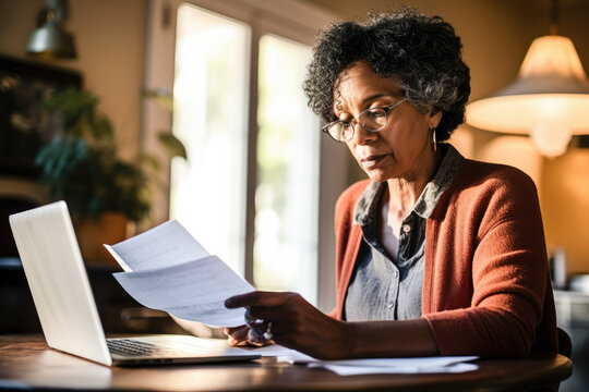 Senior woman retired sit with a laptop and reading a document. Pensive older woman with a paper bill managing her finances, planning banking loan debt, and paying taxes online using a computer. 
