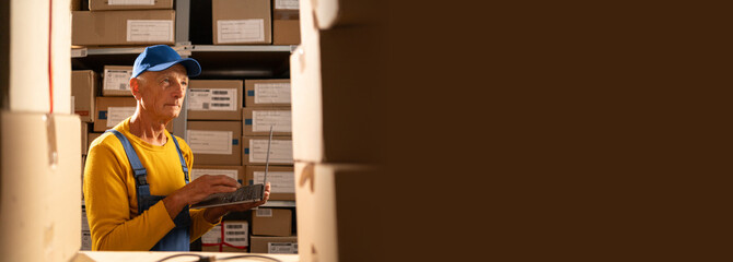 Uniformed senior worker uses laptop while standing near shelves in the warehouse.