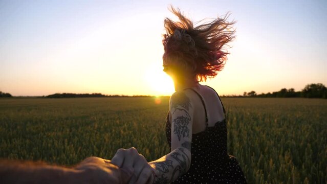 Follow me shot of happy punk girl holds male hand jogging through green wheat field at sunset. Cheerful female hipster pulling her boyfriend among barley meadow. Happy couple having fun at nature. POV