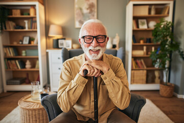 Old disabled man holding hands on wooden stick, resting in cozy armchair in living room