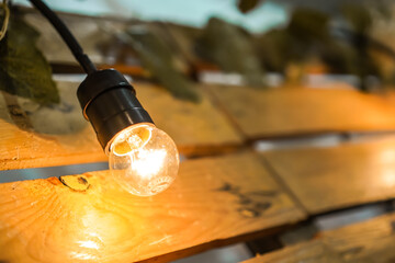 Hanging lightbulbs connected by a cable wooden planks wood background. Close-up light decoration with artificial flowers background for wedding party. Festive lights garland.