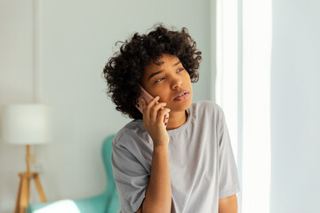 Fototapeta na wymiar African american girl talking on smartphone at home indoor. Young woman with cell phone chatting with friends. Smiling teen girl making answering call by cellphone. Lady having conversation by mobile