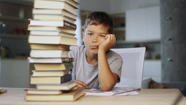 Tired exhausted stressed little boy student looking from behind large pile of books and looking at camera with sad face while preparing for exams at home Overwork education concept
