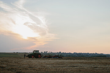 Fototapeta premium A modern tractor with a heavy trailed disc harrow plows a wheat field at sunset.