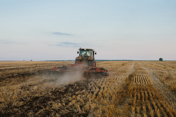 Naklejka premium Modern tractor with a heavy trailed disc harrow works a wide hilly field. Autumn or spring agricultural campaign