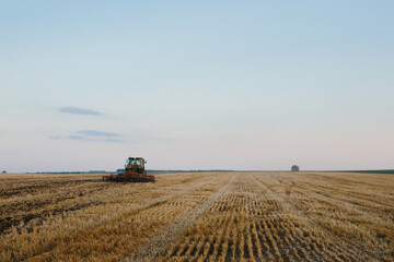 Naklejka premium Modern tractor with a heavy trailed disc harrow works a wide hilly field. Autumn or spring agricultural campaign