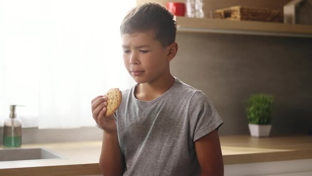 Portrait of cute little boy child eating cookie and having toothache standing at kitchen at home Sad kid feeling pain in tooth because caries gum disease or sensitive teeth indoors