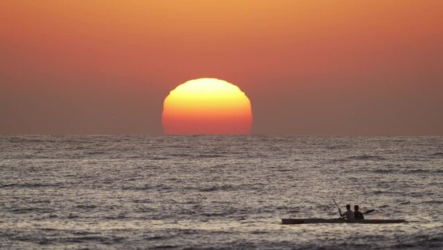 A huge, glowing sun is rising over the sea in Durban, with canoeists rowing past through the rolling waves in the foreground.