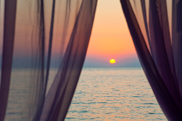 Sunset on a calm sea behind the window curtains.