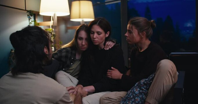 Three girls and a guy communicate in the evening in a cozy room. A sad girl talks about her experiences, and her friends support her