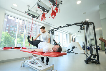 Man performing exercise with suspended straps assisted by gym instructor