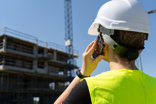 Female engineer at construction site of a building. Woman supervisor talking on the phone. Professional architect businesswoman wearing hard hat safety helmet and reflective clothing vest.