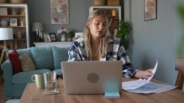 Young freelance business woman small new company owner working at home office on laptop computer drinking coffee looking financial reports stock market graphics, making decisions for clients